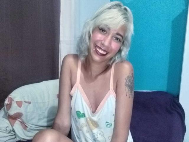 Brunna Pacheco, avec ses cheveux blonds platine, souriante, portant un caraco blanc à motifs de cœurs et bordé de rose, assise sur un lit, montrant un tatouage sur son bras droit, avec du rouge à lèvres foncé..