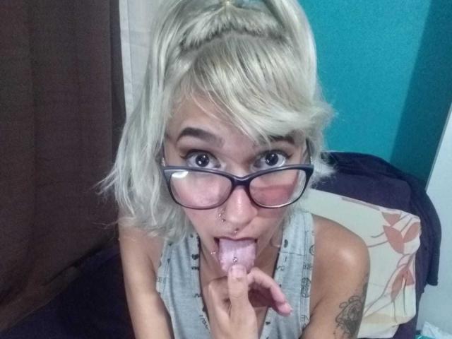 Brunna Pacheco mit blonden Haaren, Brille, Nasenring und Tätowierung, die ihre Zunge herausstreckt und ihre pinke Zungenspaltpiercing zeigt, trägt ein graues Oberteil und sitzt auf einem Bett..