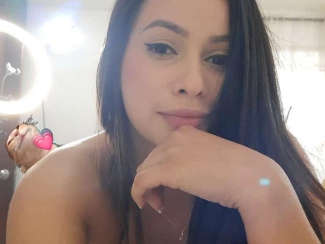 EmilyParkerr with long brown hair, wearing no clothes, resting her chin on her hand, showing subtle makeup and a cute pink heart sticker on her cheek.
