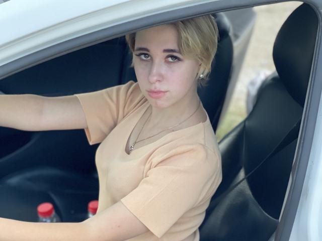 KisuLayer, con i capelli biondi, indossa una maglietta beige, è seduta in un'auto, stringe il volante, guarda in avanti con un leggero sorriso, una luce soffusa illumina il suo viso..