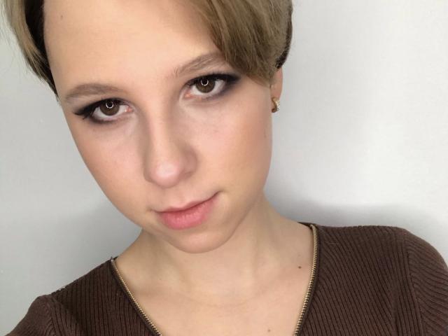 Das Gesicht von KisuLayer: kurze, braune Haare, kleine Nase, braune Augen mit dunkler Eyeliner, ein leichtes Lächeln, trägt ein braunes Rippstrickoberteil, ein kleiner goldener Ohrring..