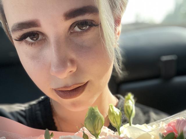 Het gezicht van KisuLayer, met bleke huid, donkere wenkbrauwen en lange wimpers, terwijl ze roze bloemen met groene knoppen vasthoudt, een donkere lipstick draagt en een zwarte top aanheeft, in een auto in het zonlicht..