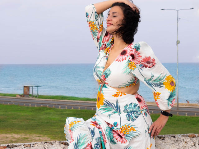 MaraRoberttss mit lockigem, schwarzem Haar, trägt ein weißes Kleid mit Blumenmuster, sitzt auf einer Steinkante, eine Hand im Haar, im Hintergrund das blaue Meer und ein bewölkter Himmel, lächelnd, kurvig, süß..
