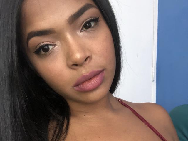 VickyGreys has smooth brown skin, thick black eyebrows, long eyelashes, and pinkish-purple lipstick. She's looking slightly to the side with straight black hair, wearing a red top.
