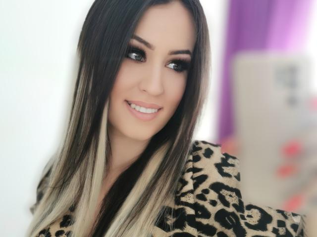 LovelyMichellex69 con cabello largo, liso, de color marrón oscuro y con un efecto degradado rubio, vistiendo un abrigo de estampado de leopardo, sonriendo a un teléfono inteligente, sosteniendo una botella beige y con uñas pintadas de rosa..