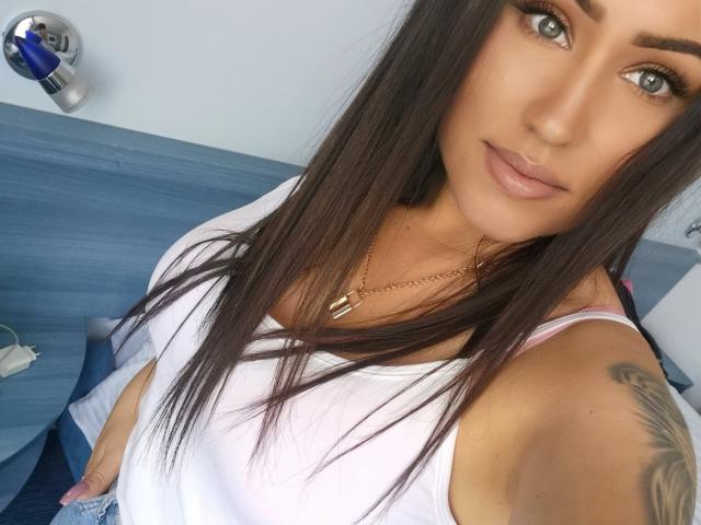 LovelyMichellex69 with long brown hair, green eyes, and a subtle smile, wearing a white tank top, showing a tattoo on her right arm, and a gold necklace.