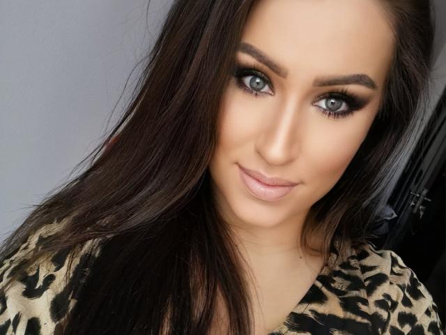 A LovelyMichellex69 tem olhos azuis, cabelo castanho escuro e uma maquiagem com olhos esfumados e cílios grossos. Ela está vestindo uma blusa com estampa de leopardo e sorri suavemente..