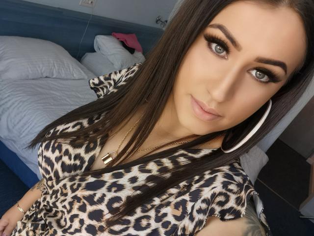 LovelyMichellex69, con cabello castaño largo, ojos verdes y una blusa de estampado de leopardo, lleva grandes aros y un collar de oro, y tiene un tatuaje en su brazo derecho..