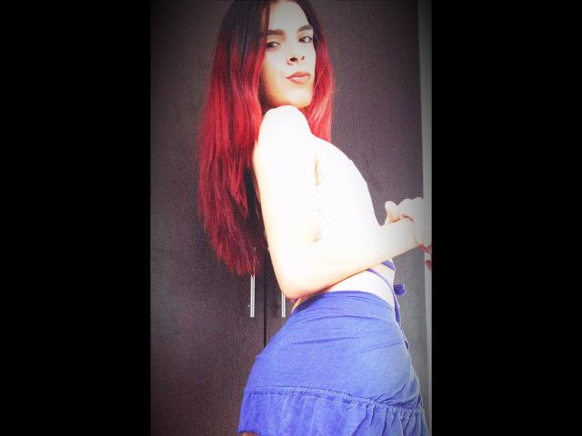 KataleyaHoty with red hair, topless, showing her ass in a blue skirt, standing sideways, looking back, smiling, hands near waist, bright sunlight, black background.