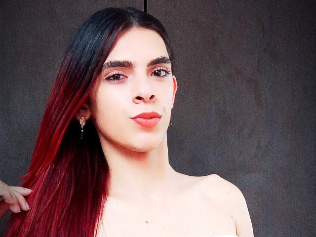 KataleyaHoty has long red and black gradient hair, fair skin, red lipstick, small earrings, and a neutral expression. She's topless with a slight smile.