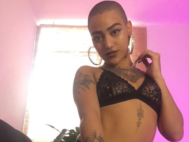 RoseFromHell mit raspelkurzem Kopf, trägt einen schwarzen Spitzen-BH, große Ohrringe, Tattoos auf Brust und Arm, hält einen Ohrring, rosafarbenes Licht, blickt in die Kamera, leichtes Grinsen..