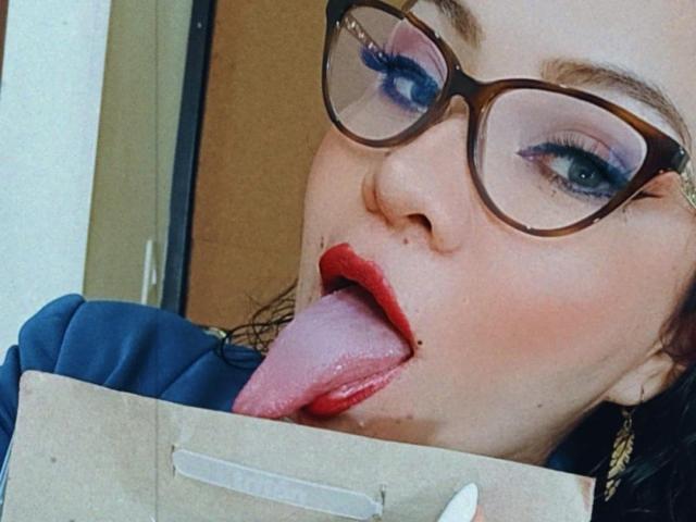 AlisGourmande with red lipstick, blue eyeshadow, and glasses, tongue out, licking a brown paper bag, showing a small cut on her tongue. Her tits are partially visible.