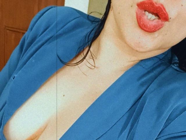 AlisGourmande with red lips, blue shirt, and visible cleavage, showing a bit of her tits. Her black hair is partially visible, and she has a small mole on her neck.