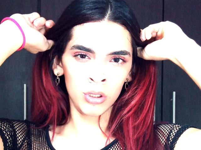 KataleyaHoty tem cabelo ombré vermelho, blusa de malha preta, pulseira rosa, segurando o cabelo, olhos castanhos, lábios levemente entreabertos, pequenos brincos de argola, pele clara, olhando para a câmera..