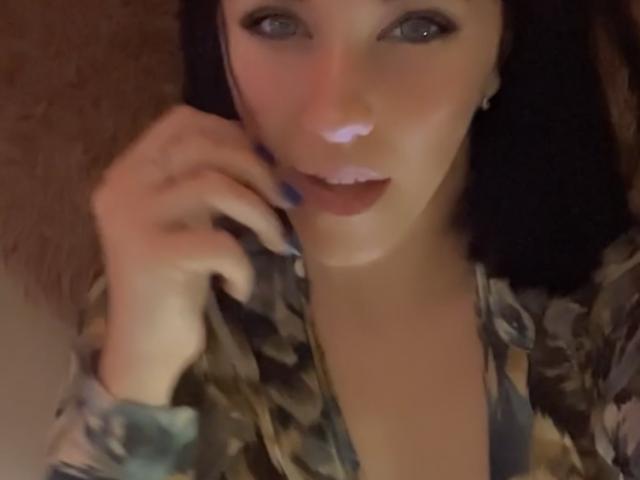 Misstyk with dark hair, blue eyeliner, and a pink nose, wearing a camo jacket, has blue nail polish, touching her cheek, looking at the camera.