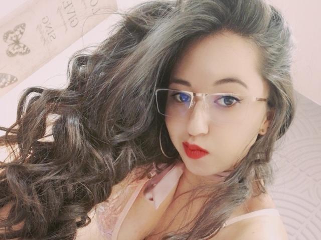 IsabellaWang with long, wavy black hair, wearing glasses, red lipstick, and a pink lace bra, showing off her tits in a selfie. Her blue eyes are looking at the camera.
