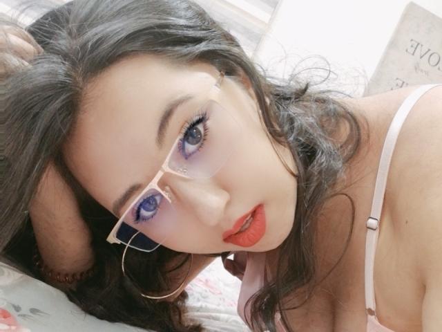 IsabellaWang with pink glasses, red lipstick, and hoop earrings, lying on a bed, looking up with blue eyes, white bra straps visible, dark wavy hair, and a slightly open mouth.