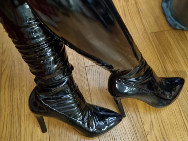 Les bottes en latex noir brillantes de CocoMelle, avec des talons hauts et ajustées autour de ses cuisses, se reflètent sur le plancher en bois. Elle est debout, les jambes légèrement écartées, les bottes pointées vers l'avant..
