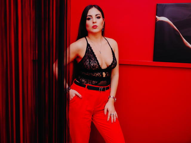BelleInconnu has big boobs in a black lace top, red pants, and long dark hair, leaning against a red wall, with a sexy butt photo on the right.