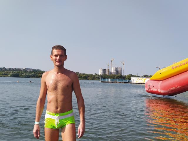 PierceBrick se tient dans l'eau, torse nu, portant un maillot de bain vert néon et blanc, avec un bateau gonflable rouge et jaune sur sa droite. Il a une poitrine musclée et poilue..