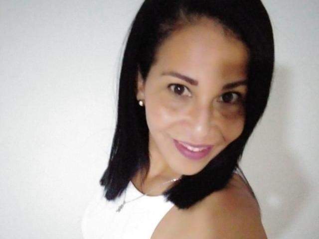 Shanon, una donna matura con capelli neri lunghi fino alle spalle, occhi marroni e una camicetta bianca, sorride alla fotocamera, mostrando il suo rossetto rosa e piccoli orecchini di perle..