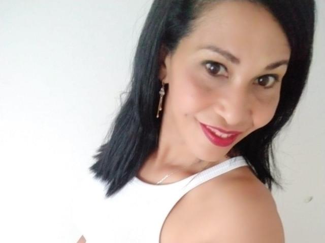 Shanon, volwassen, met een medium bruine huid, zwart haar tot op de schouders, rode lipstick, een wit tanktopje en oorbellen, lacht naar de camera. De borsten zijn zichtbaar onder het tanktopje..