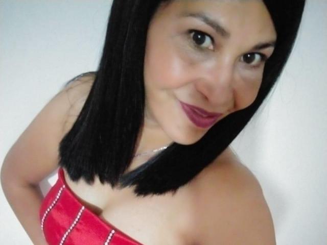 ShanonMature with black straight hair, red strapless dress, and red lipstick, smiling at the camera, showing her boobs, small silver necklace, light skin, and brown eyes.