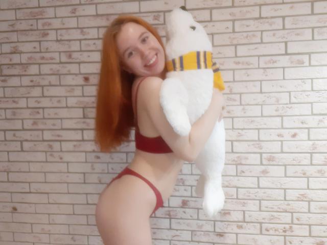 AnikaGlow with red hair, smiling, wearing red bra and panties, holding white stuffed bear in yellow striped shirt, her ass and tits visible, against white brick wall.