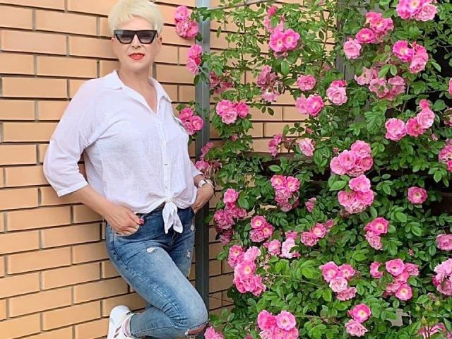 NikaClare met kort blond haar, een zonnebril op, een wit overhemd met knopen dat vooraan dichtgebonden is, blauwe jeans en witte sneakers, staat naast roze rozenstruiken..