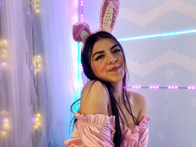 MiaDupont com orelhas de coelho rosa e brancas, cabelo preto longo, blusa rosa com um ombro descoberto, sorrindo, bochechas coradas e um piercing nasal fofo, parecendo sexy com uma expressão divertida..