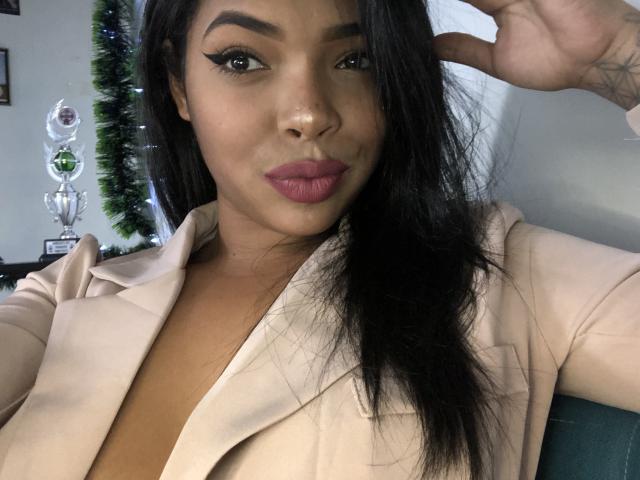 VickyGreys with long black hair, pink lipstick, and a beige blazer, making a peace sign, showing her tits slightly, looking at the camera with a cute smile.