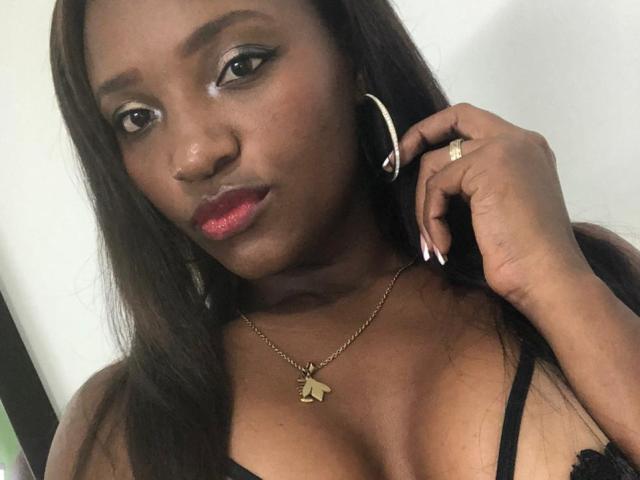 GiceleSweety with dark skin, long brown hair, wearing a black lace top, hoop earrings, gold necklace with butterfly pendant, red lipstick, and a ring, looking at camera.