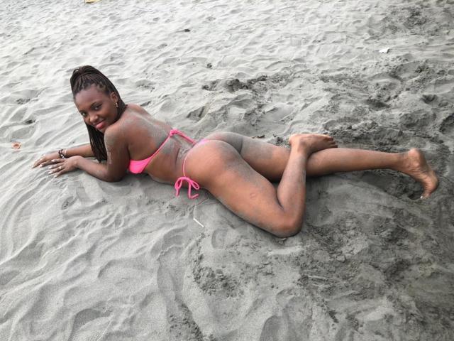 GiceleSweety está acostada boca abajo, con un bikini rosa, mostrando su trasero y la hendidura entre sus glúteos, con arena en su cuerpo y una sonrisa en su rostro..