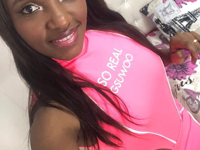 GiceleSweety, avec sa peau foncée, ses longs cheveux noirs et son rouge à lèvres rose, sourit à l'appareil photo. Elle porte un haut rose vif à col haut avec du texte blanc, qui révèle sa poitrine..