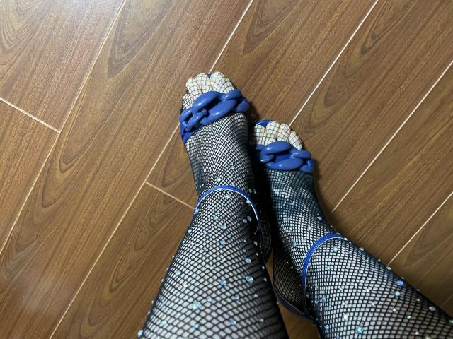 Les pieds d'AndraD portent des bas de pêche noirs avec des nœuds bleus et de petits clous bleus, et sont posés sur un plancher en bois. Les orteils sont légèrement recourbés..