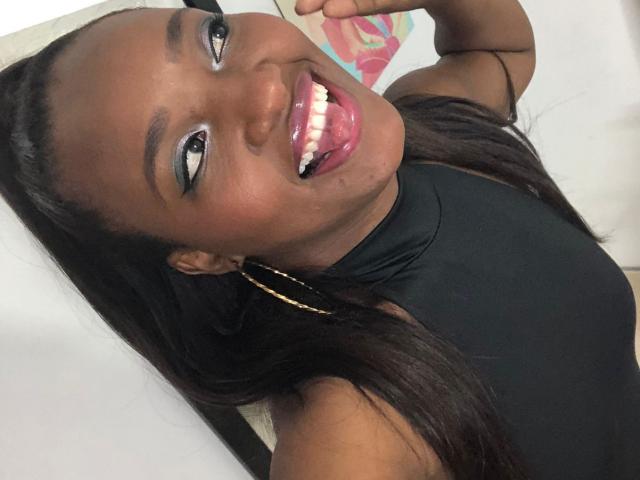 GiceleSweety, con la pelle scura, una canottiera nera, orecchini a cerchio dorati e lunghi capelli neri, che mostra la lingua rosa, sorride mostrando i denti bianchi e ha il trucco azzurro sugli occhi..