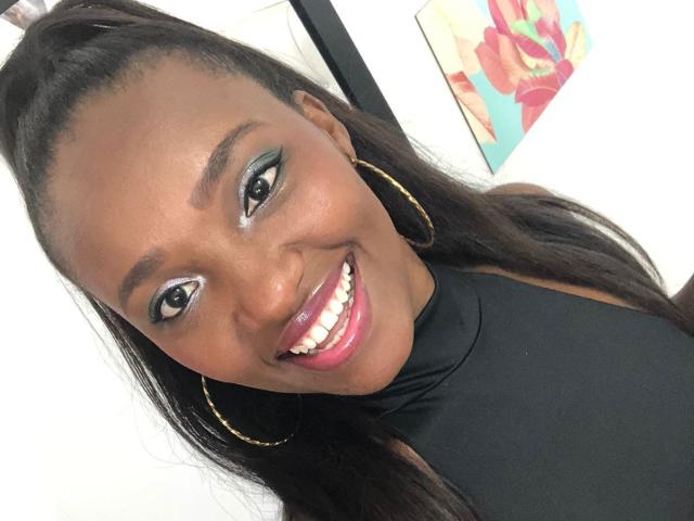 GiceleSweety with dark skin, long black hair in a ponytail, gold hoop earrings, black top, shiny pink lips, and teal eye makeup, smiling at the camera.