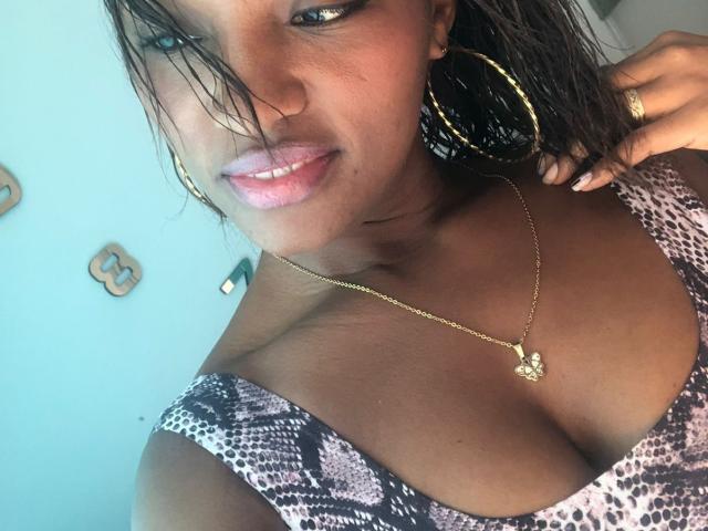GiceleSweety, con la pelle marrone scura, i capelli neri bagnati, orecchini a cerchio dorati, una collana dorata, rossetto rosa e un vestito a stampa leopardo che mette in mostra il suo seno grande e lucido..