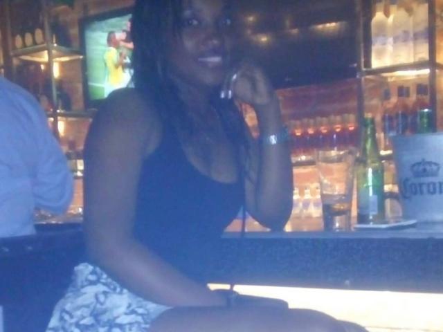 GiceleSweety, à la peau foncée et aux longues tresses, portant un débardeur noir et un short blanc à motifs, est assise au bar, parlant au téléphone, avec une bouteille et un verre à proximité..