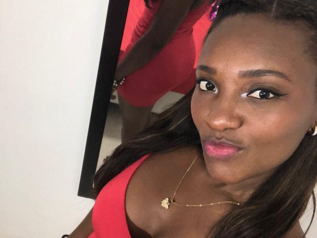 GiceleSweety with dark skin, pink lipstick, and gold necklace, wearing a red top, showing her tits in a mirror selfie, her brown hair in braids, looking at the camera.