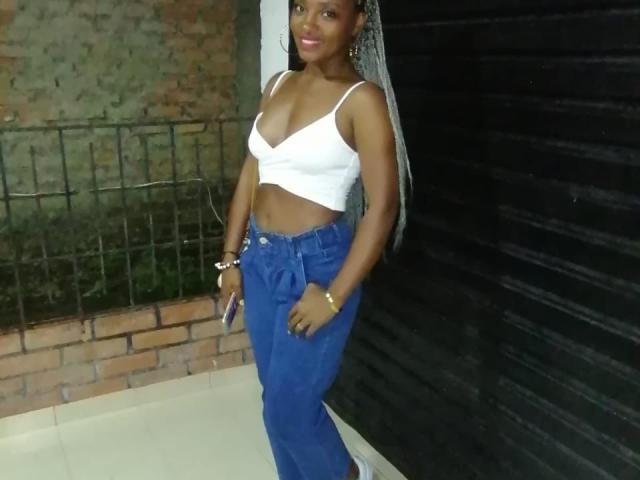 GiceleSweety with dark skin, long braided hair, white crop top showing her tits, blue jeans, holding a phone, standing against a black door, wearing a gold bracelet.