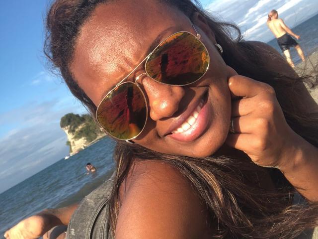GiceleSweety, avec une peau brun foncé, de longs cheveux ondulés, souriante, portant des lunettes de soleil aviateur dorées réfléchissantes, montrant un nez mignon et un large sourire blanc. Sa main est sur sa joue..