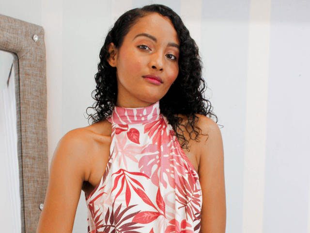 Megan Gil, avec ses cheveux noirs bouclés, porte une robe à bretelles à fleurs rose et blanche, qui révèle ses petites seins, et affiche un sourire subtil et confiant. Elle a un teint brun clair..