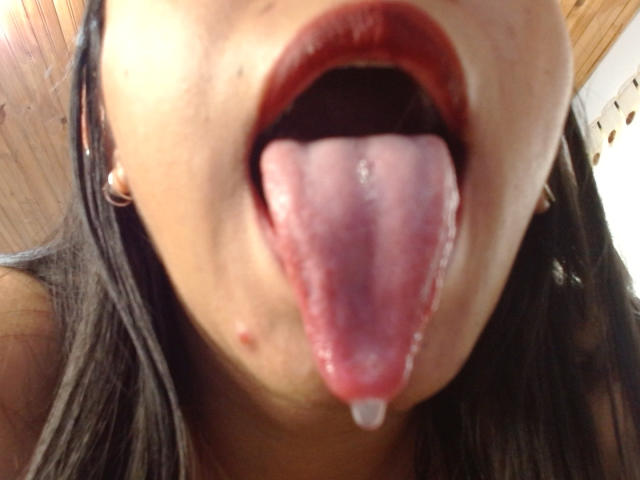 La bocca di AshleyLover è spalancata, con la lingua che le sporge fuori, ricoperta da una piccola goccia di saliva, e indossa un rossetto rosso. I suoi capelli castani sono parzialmente visibili..