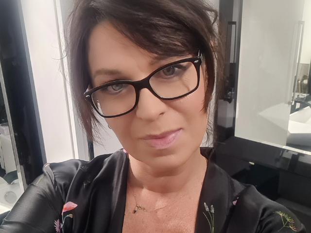 KellyMatureX heeft kort bruin haar, een zwarte bril en draagt een zwarte jurk met bloemenprint. Haar roze lippen en subtiele make-up zijn opvallend. Ze heeft een gouden ketting..