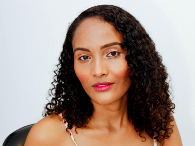 MeganGil a les cheveux noirs et bouclés, les yeux marron et du rouge à lèvres rose. Elle porte un haut rose et blanc, avec un sourire discret, et a un visage lisse et de couleur marron..