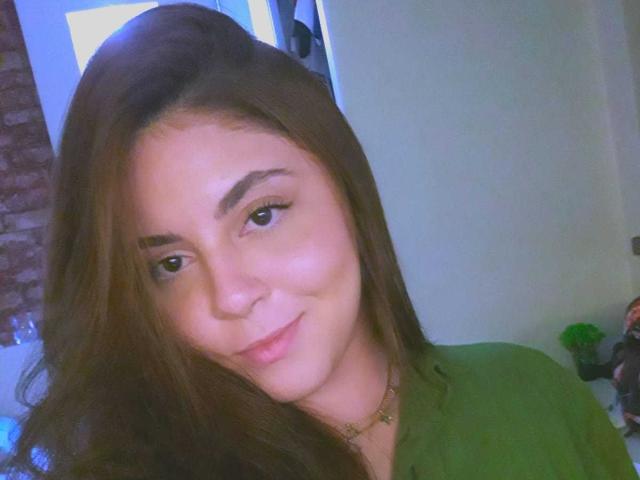 Anynane tem cabelos castanhos longos, olhos castanhos e um sorriso discreto. Ela está vestindo uma camisa verde e um colar dourado. Suas nádegas não estão visíveis. Seus seios não estão visíveis..