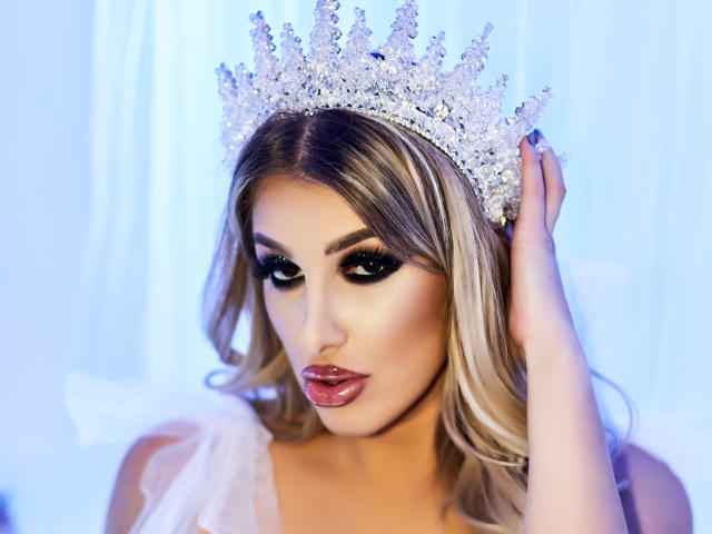 TanyaMonroe with a sparkly silver tiara, blonde hair, and dark makeup, touching her head with her right hand, wearing a sheer white top, looking seductive.