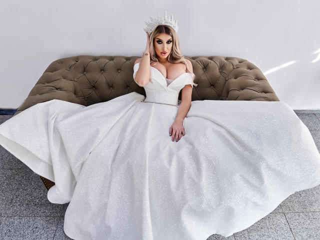 TanyaMonroe with big tits, wearing a sparkling white dress and a crown, sitting on a brown tufted sofa, hand on her head, looking at the camera.