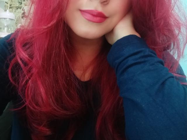AhriValvet, com cabelos ruivos vibrantes e ondulados, vestindo uma camisa azul escura de manga comprida, usa batom vermelho e apoia a cabeça na mão. Seus seios estão parcialmente visíveis..